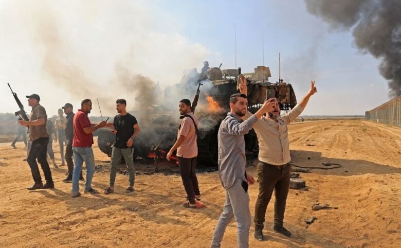 Men with guns stand in front of a burning tank.