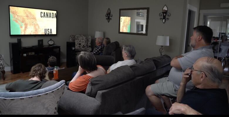 A group of people sit in a living room, watching a documentary film on a flat-screen television.