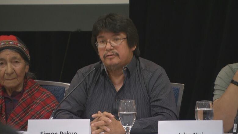 A man wearing a black shirt sits at a table in front of a microphone.