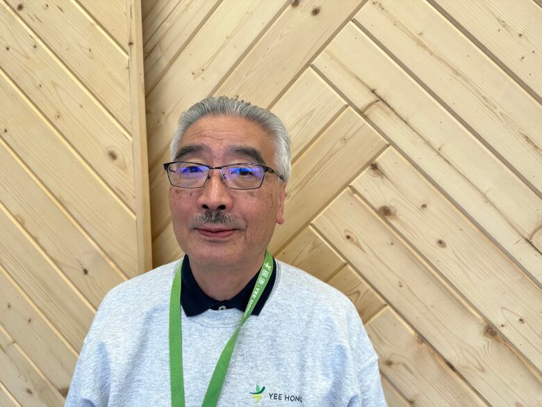 A man with grey hair and glasses stands against a wooden wall and smiles slightly. 