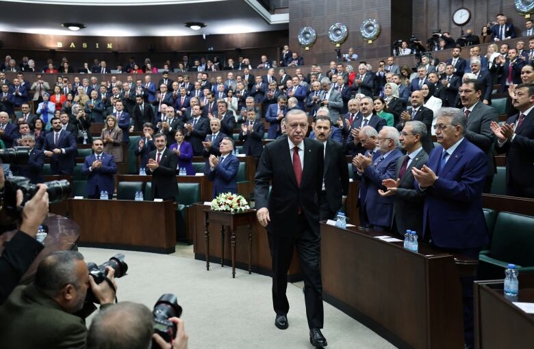 A man in a suit and tie walks as dozens of people stand and applaud in a rounded chamber.