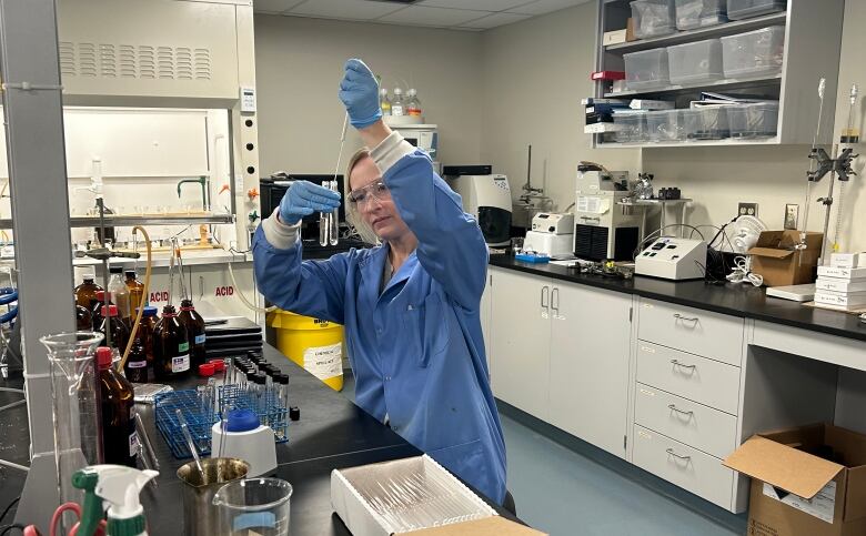 A woman in a blue lab coat holds a beaker and a syringe, with lab equipment in the background.