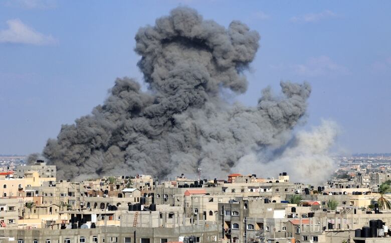 Large clouds of dark smoke fill up the blue sky above a city filled with low-rise buildings.