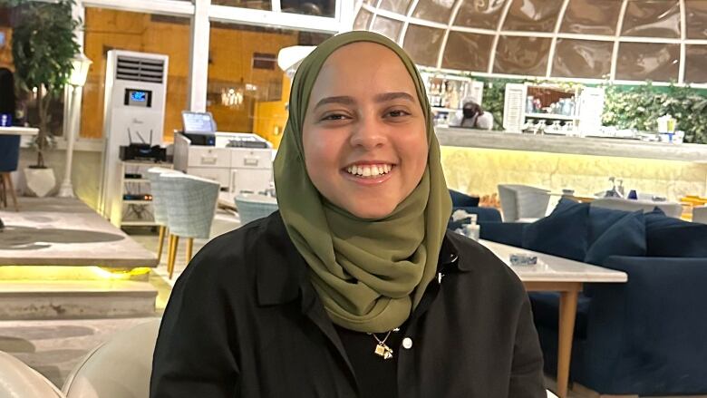 A woman in a green head covering sits at an indoor cafe. She is smiling at the camera.