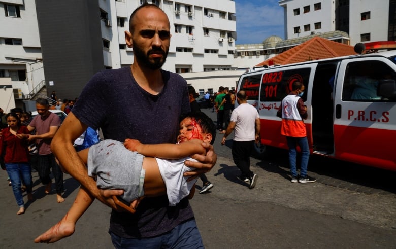 A man carrying an injured child, with an ambulance van in the backdrop. 