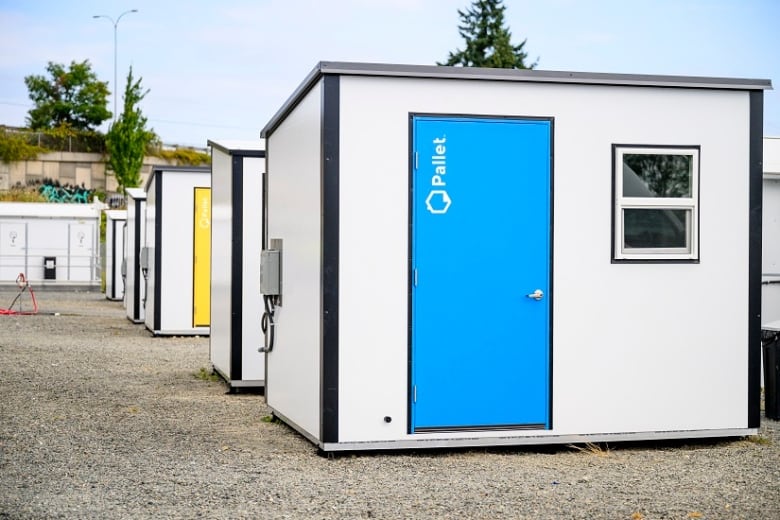 A small white structure with a blue door sits at the front of a row of a identical structures, one of which has a yellow door.