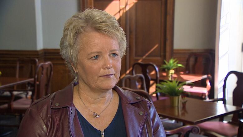 A woman with a sombre face and short blonde hair looks off camera. She wears a burgundy leather coat.  