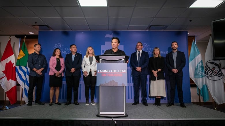 An East Asian man stands at a podium titled 'Taking the lead on housing', flanked by councillors on either side.