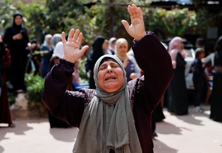 A woman in headscarf holds her hands aloft with an expression of grief in an outdoor photo.