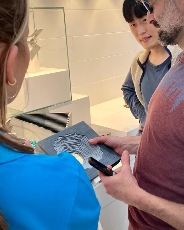 A man holds a small, gray Lego structure and shows it to two onlookers.