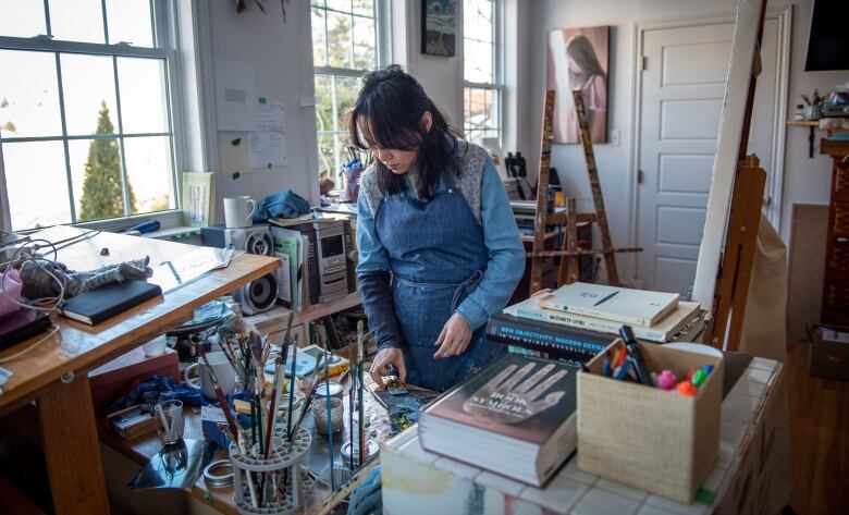 A woman in a blue shirt and grey vest with a blue apron stands in front of a table covered with art supplies.