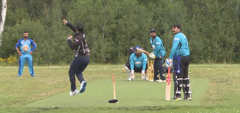 A man takes a flying, wind-up pitch down a green section of grass toward a man with a flat bat, while other men stand around near the batter.