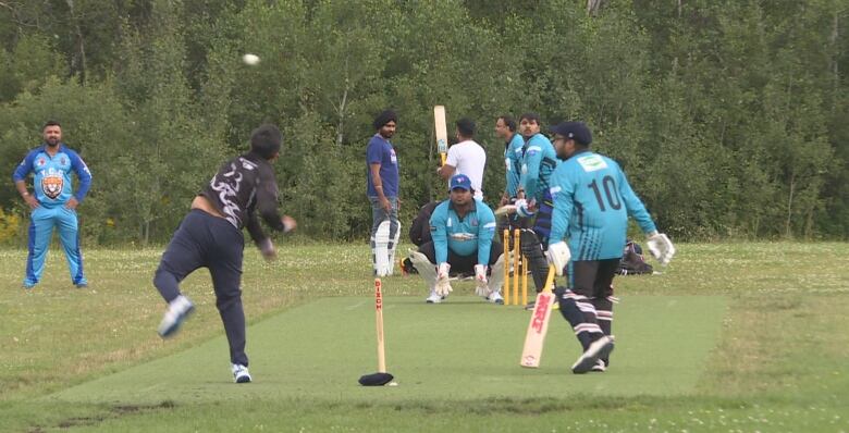 A man takes a flying, wind-up pitch down a green section of grass toward a man with a flat bat, while other men stand around near the batter.