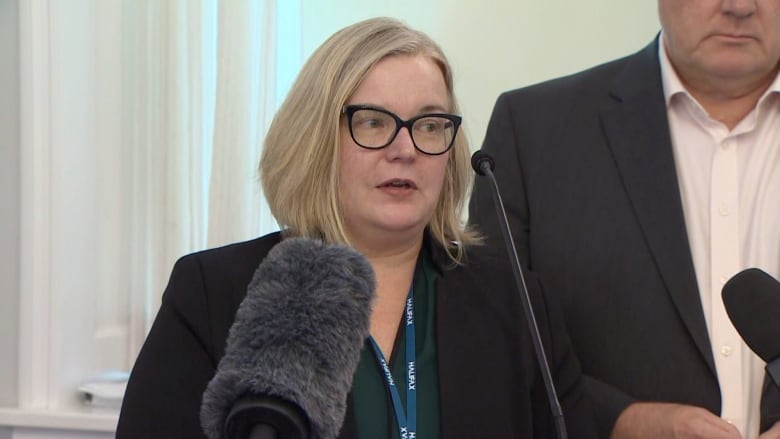 A white woman with glasses and blonde hair in a bob stands at a podium with microphones in front of her