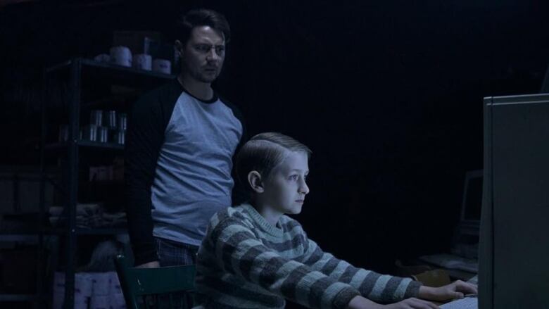 A man stands behind a boy sitting at a computer. The man appears frightened. 