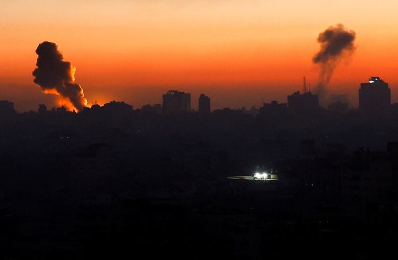 Two plumes of smoke are silhouetted against an orange sky. One sole, bright light is in the foreground.