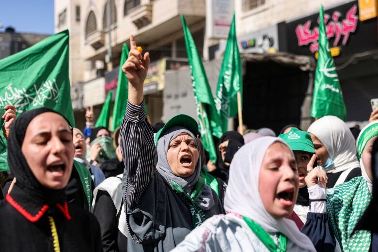 Several women in headscarfs appear to be yelling and chanting in an outdoor demonstration, with many holding up flags.