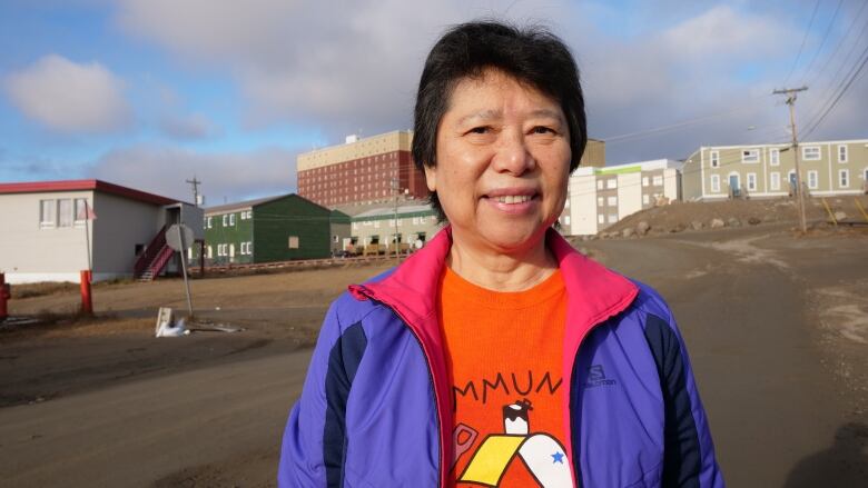 A woman stands outside with an orange shirt and purple jacket with a brown dirt road in the background. 