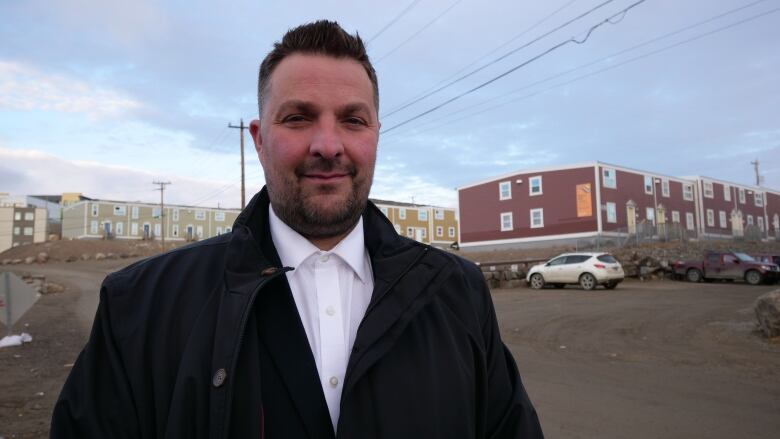 A man wearing a white collared shirt and a black jacket looks at the camera with a brown dirt road behind him. 