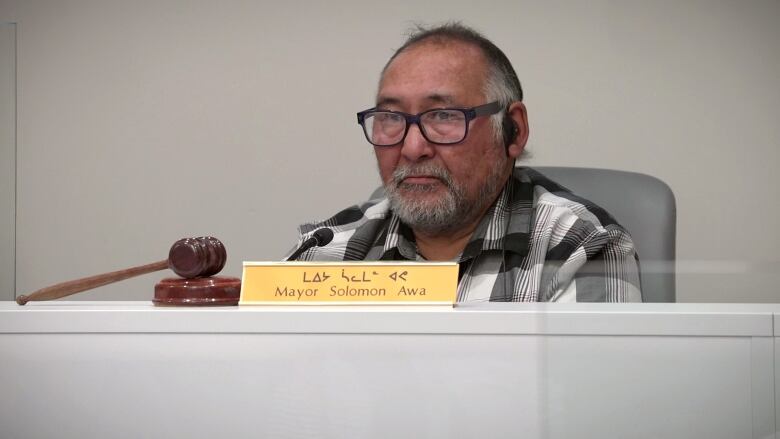 A man with glasses sits at a desk behind a name plate with Mayor Solomon Awa written on it. 