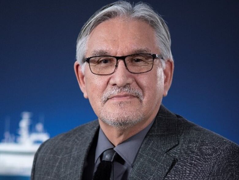 A man with glasses, grey hair and a grey beard wearing a black suit and black ties poses for a headshot. 