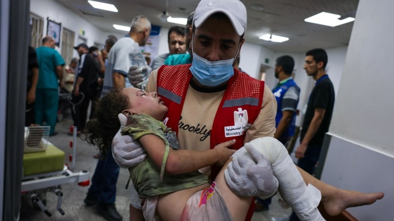 A man in a red vest carries an injured child. 