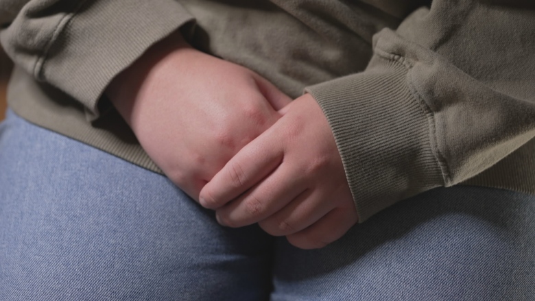 A woman's hands are folded in her lap. She is wearing jeans and a green sweatshirt.