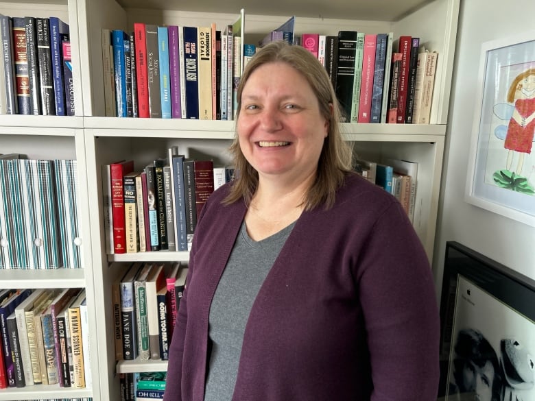 A woman stands at a slight angle to the camera, smiling. She has blonde shoulder length hair and is wearing a purple cardigan sweater over a grey top. She is standing in front of a bookshelf.