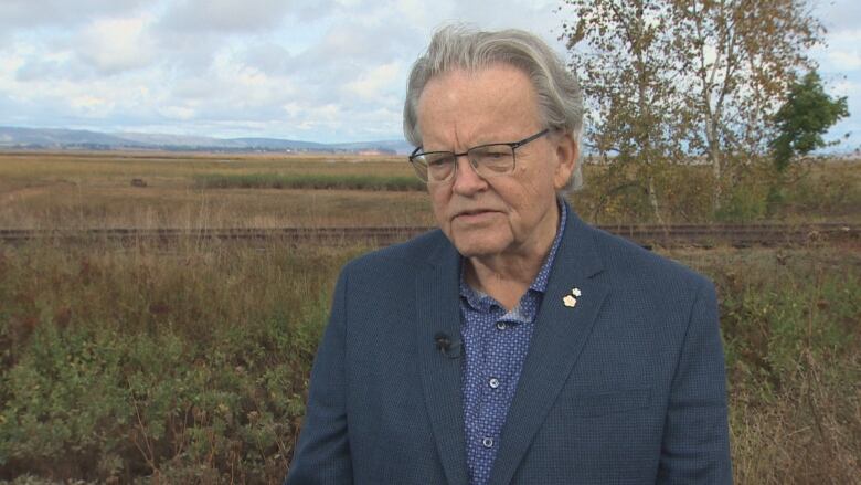 An old white man with grey hair stands in an empty field. He's wearing a dark blue checkered suit jacket over a blue button up with small dots. He has two small flower pins on his lapel.