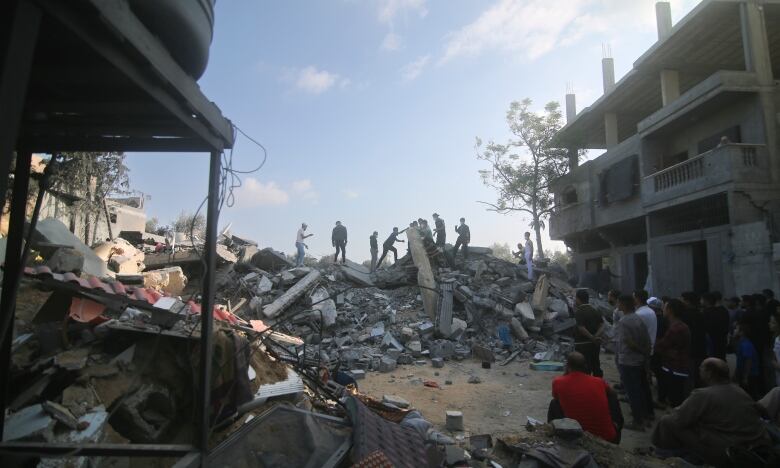 People stand on a large pile of rubble surrounded by destroyed buildings.