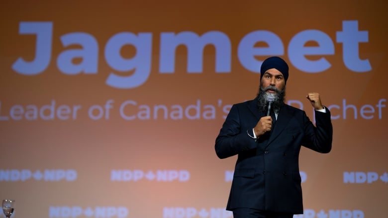 A bearded man, wearing a turban and a dark suit, gestures as he speaks into a microphone.