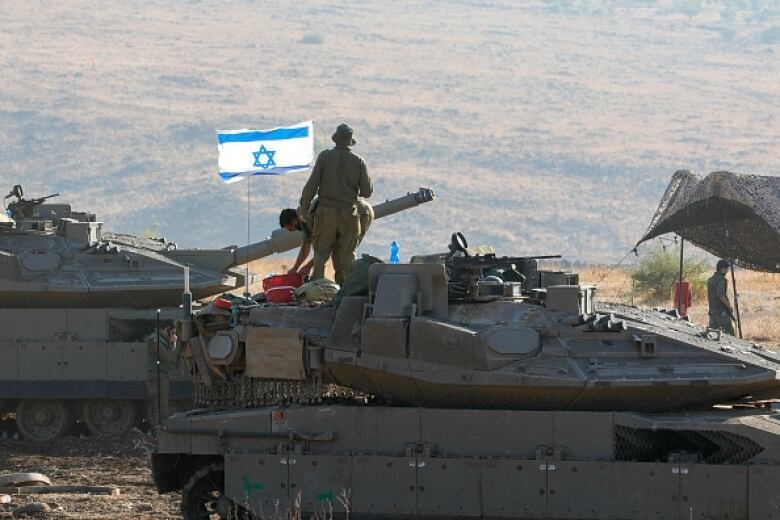 Two soldiers stand on a tank positioned overlooking a valley. 