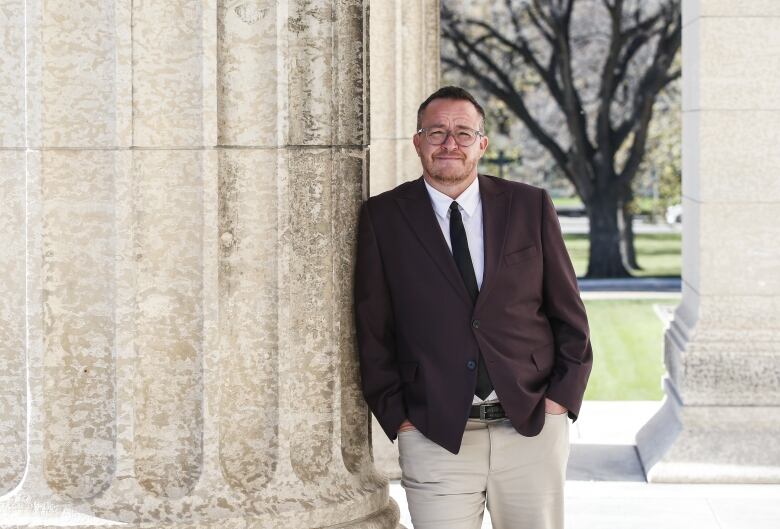 A person wearing a suit and tie leans on a column attached to a building.