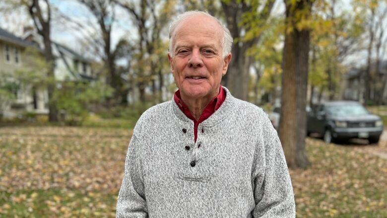 A man with short grey hair, wearing a grey sweater, stands on a sidewalk and stares into the camera.