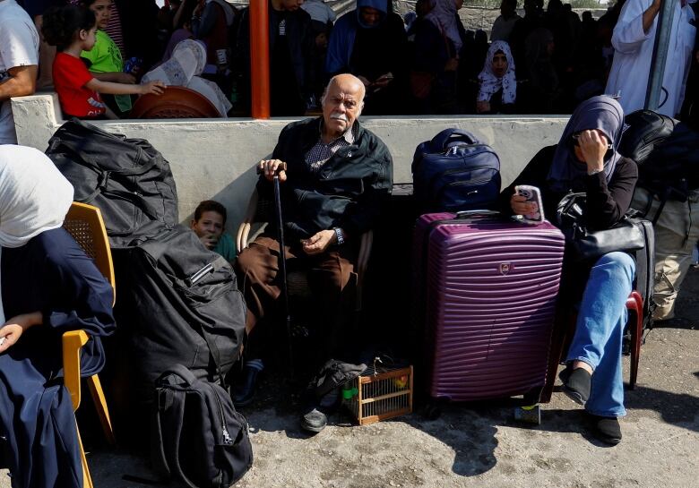 An older couple sits with their suitcases at a closed border crossing on a hot day.