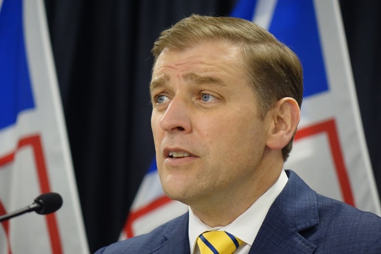 A man wearing a yellow tie looks to the left with flags in the background.