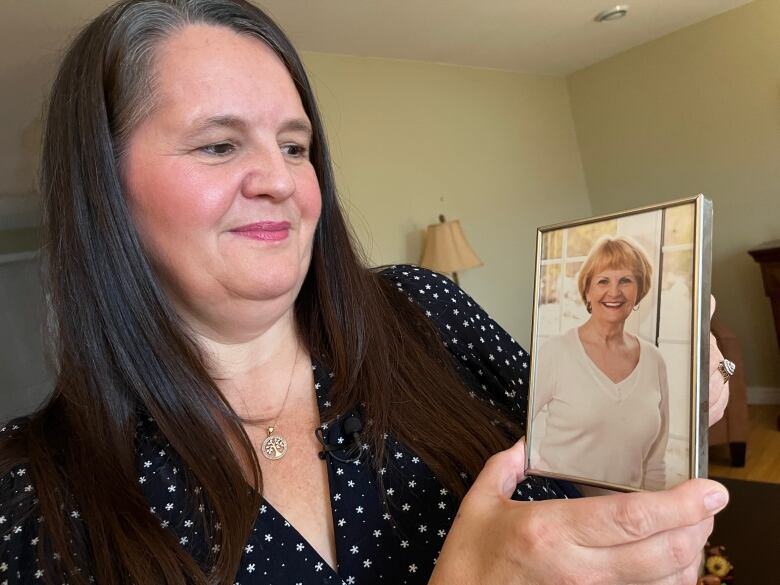 A woman with long dark hair holds a picture in a frame of an older woman in a white sweater.