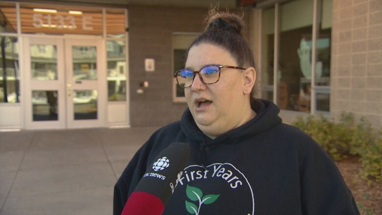 A woman in a black hoodie speaks with a reporter. A red and black CBC microphone can be seen in front of her. 