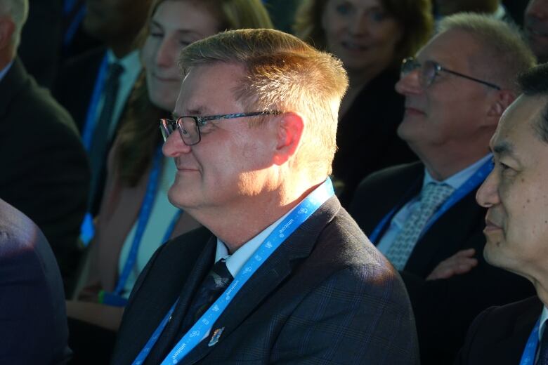 A man with neat reddish brown hair parted to the side and glasses, sits among a crowd of people wearing business suits.