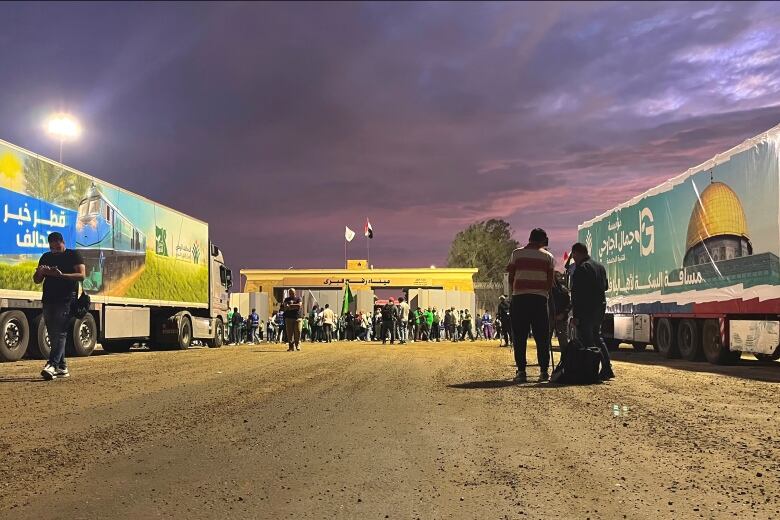 Parked tractor-trailers, part of a humanitarian convoy) face the Rafah border crossing on the Egypt side, with people milling about, early morning.