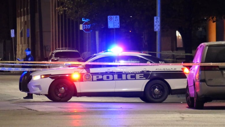 A police officer stands near a black and white cruiser with its lights on. The street in blocked with yellow tape.