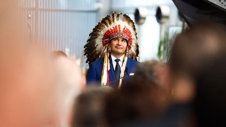 A man wearing a headdress stands in front of a crowd of people.