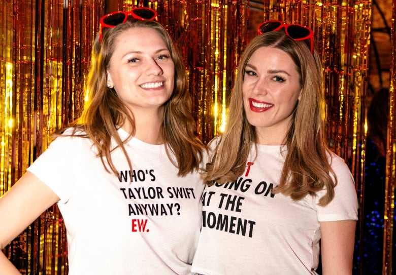 Two women with their arms around each other stand in front of a gold foil curtain.