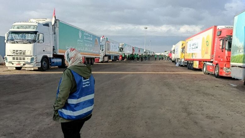 A person wearing a blue safety vest identifying them as a volunteer stands before a parked convoy of trucks