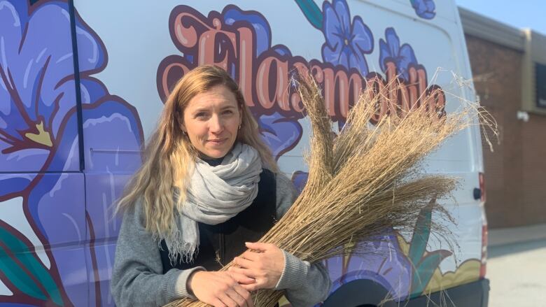 A woman holding a bundle of dried flax stalks stands in front of a cargo van.