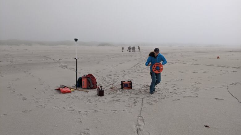 A person wearing a blue jacket stands on the sand with measuring equipment on a foggy day.