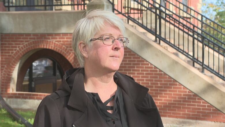 Woman in glasses with short white hair stands outdoors in front of a brick staircase.