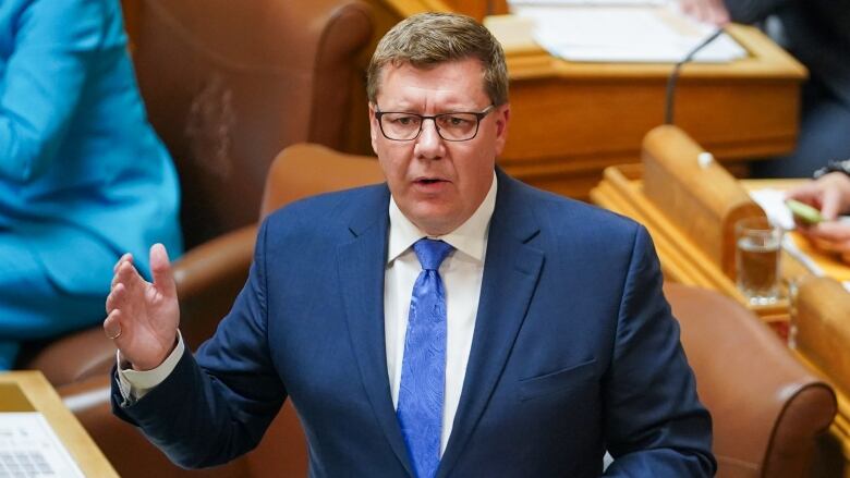 A man in a blue suit stands and speaks in a legsislative chamber.