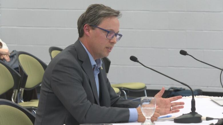 The mayor is seated speaking at a microphone. He is wearing glasses and a grey suit with a blue shirt. 