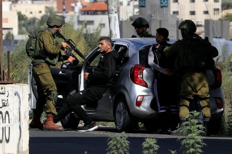 A helmeted soldier points a long gun at a man pressed against a car in an urban setting.